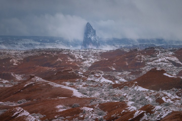 Snow-covered red rock desert with a distant pointed mountain peak under cloudy skies.