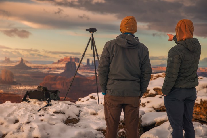 Two people in winter clothing view a snowy desert landscape with a tripod.