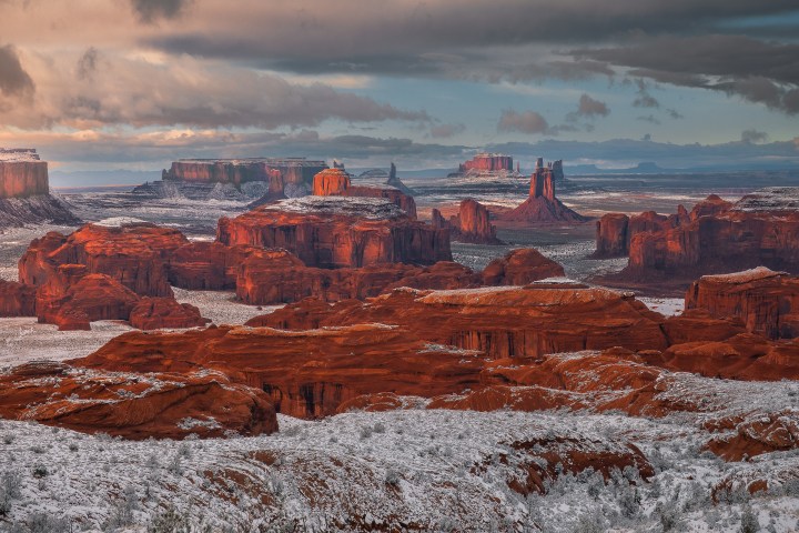 Snow-covered desert landscape with red rock formations and cloudy sky.