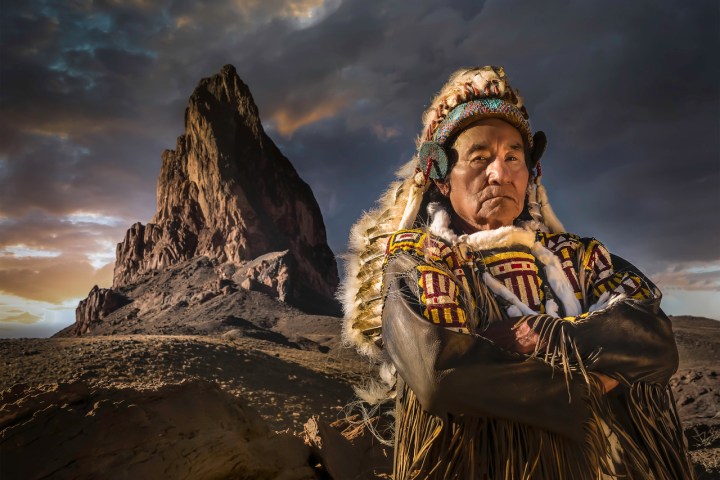 Person in traditional attire stands before a rocky mountain at sunset.
