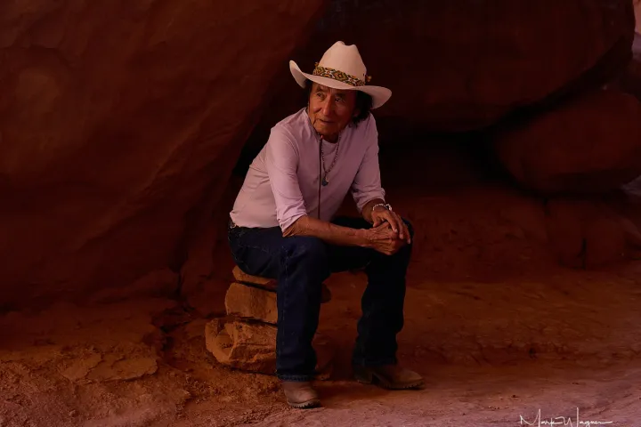 Person in white hat and shirt sitting on rock under large boulder.