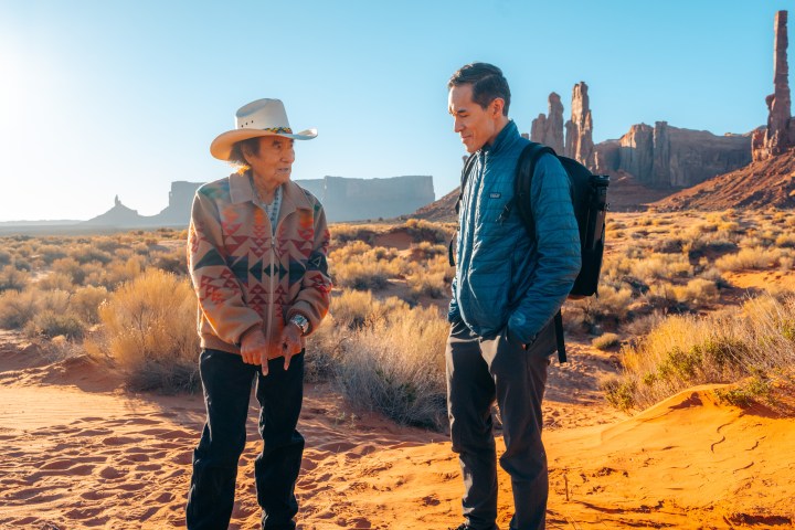 Two people talking in a desert with rock formations in the background.