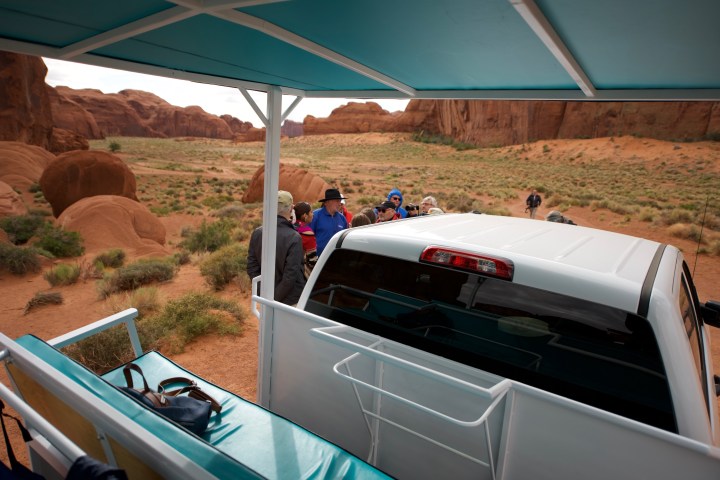 Tour group near a white vehicle in a desert landscape with red rock formations.