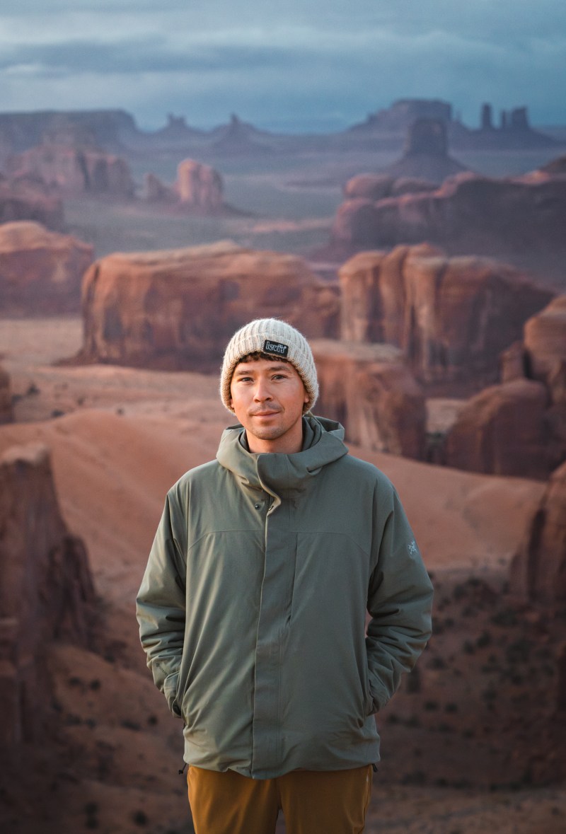 Person in a jacket and beanie stands before a canyon landscape at sunset.