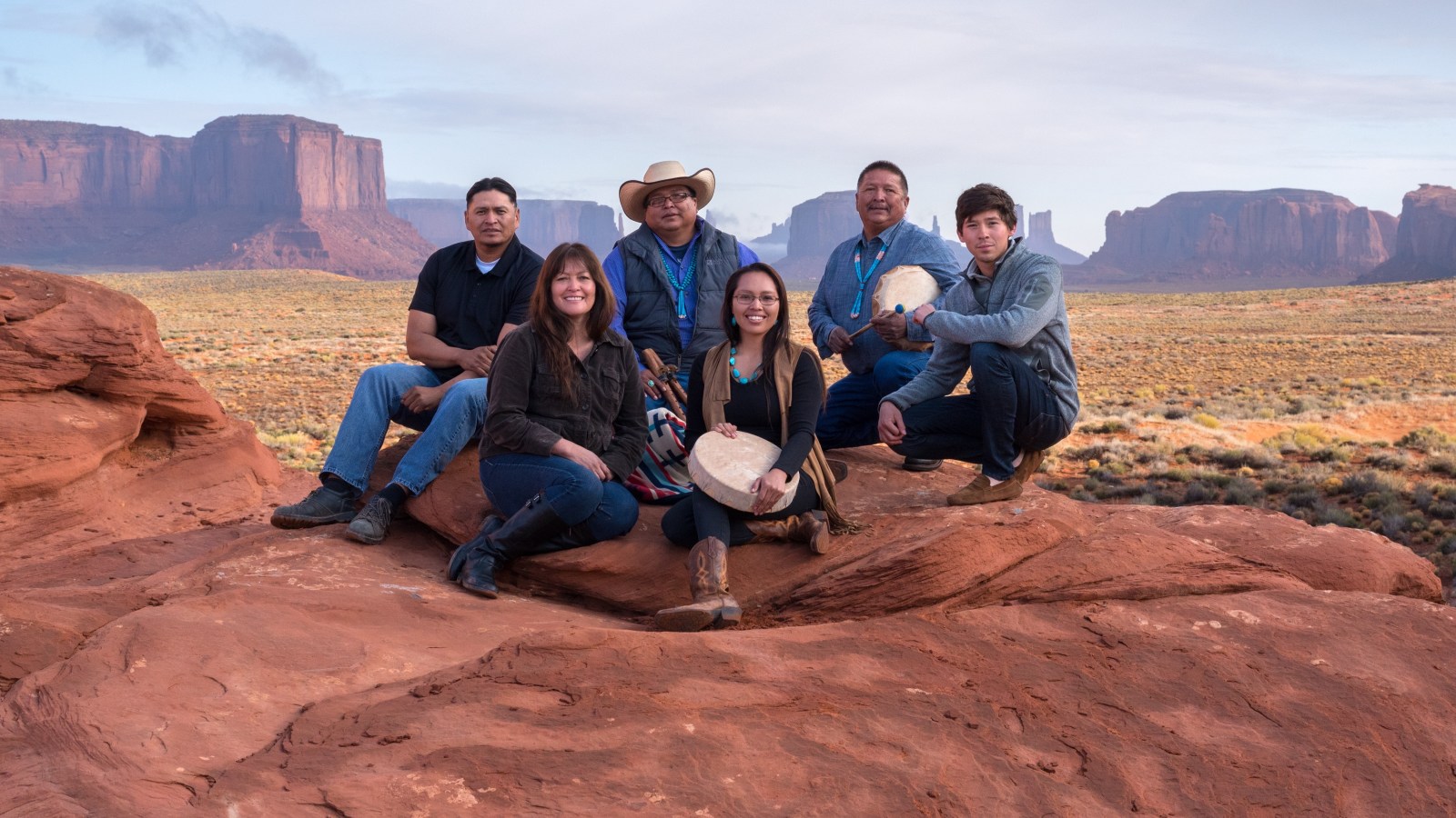 Group of six people sitting on red rocks with a desert landscape in the background.