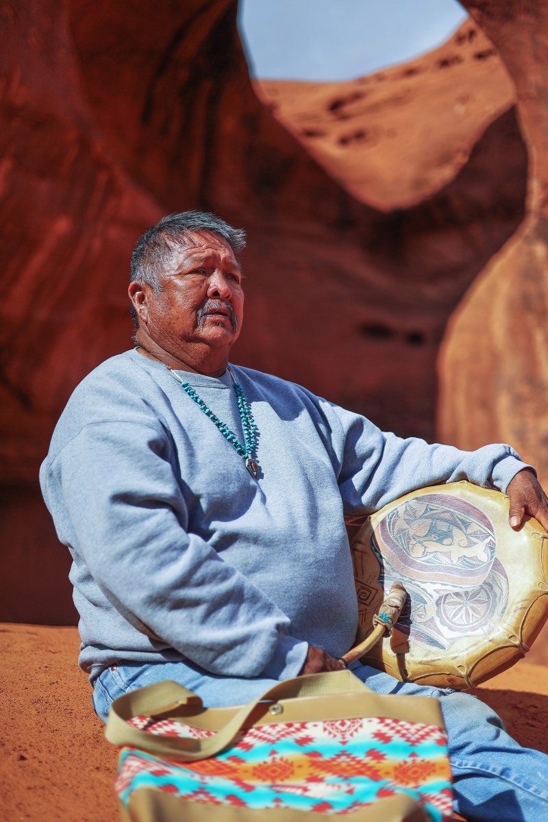 Person sitting outdoors with drum and colorful bag, rock formations in background.
