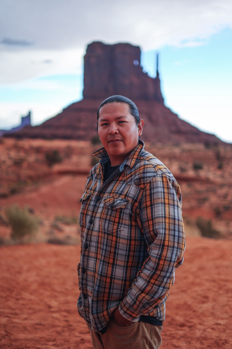 Person in plaid shirt standing in desert landscape with butte in background.