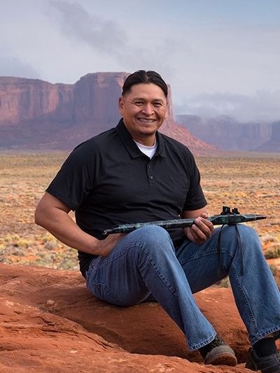 Man with a rifle sitting on red rock with desert and cliffs in background.