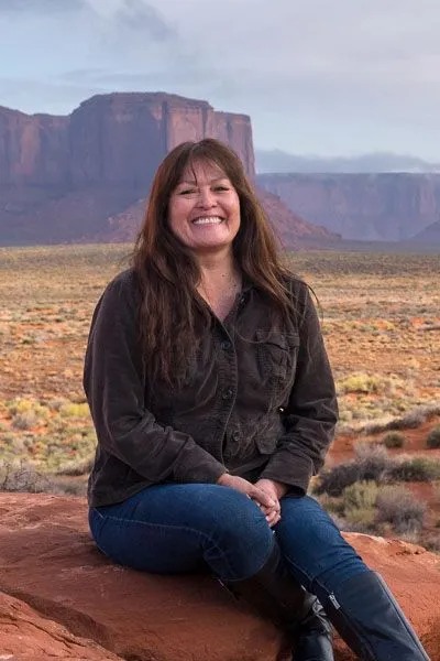 Person smiling seated on a rock with desert landscape and mesas in the background.