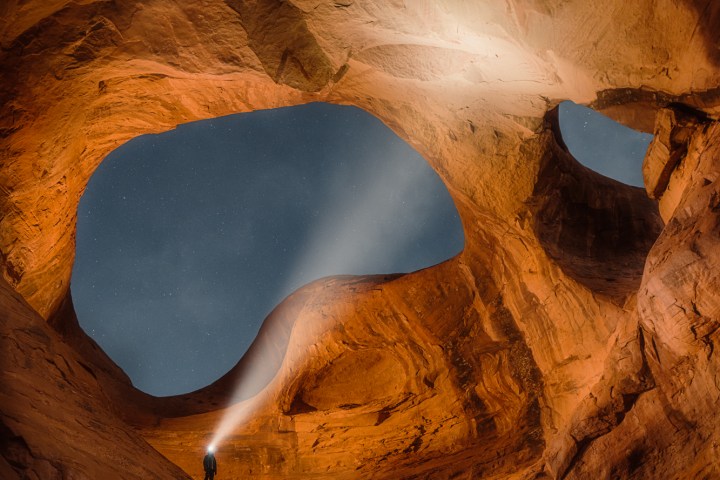 Person with flashlight in illuminated red rock cave arch at night.